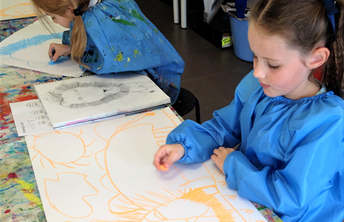 Young girl wearing a blue painting smock drawing the outline of a lion on a white canvas using yellow crayon